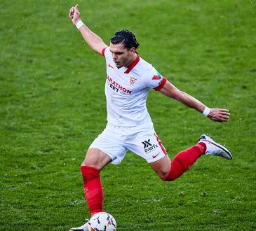 Ein schwarzhaariger Fussballspieler im weißen Trikot schießt einen Ball