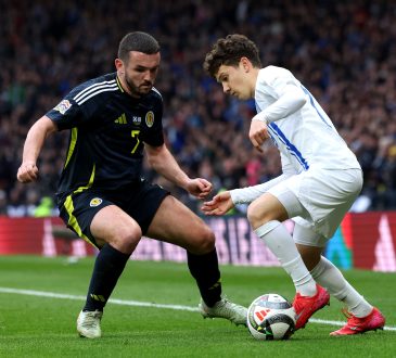 Zwei Spieler stehen sich gegenüber bei einem Fußballspiel. Der linke Spieler trägt ein schwarzes Trikot und spielt für Schottland. Rechts steht ein griechischer Nationalspieler in einem weißen Trikot. Der Griechische Spieler heißt Karetsas und versucht in diesem Moment an seinen Gegenspieler mit einem Dribbling vorbei zu kommen.