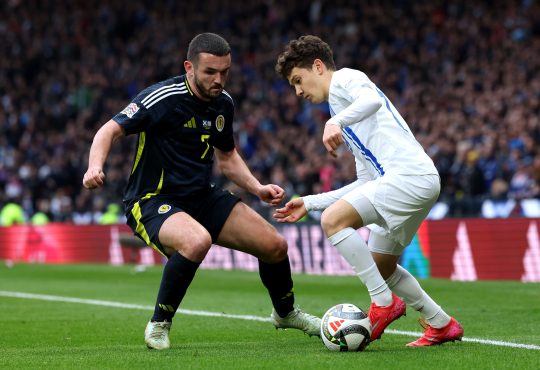 Zwei Spieler stehen sich gegenüber bei einem Fußballspiel. Der linke Spieler trägt ein schwarzes Trikot und spielt für Schottland. Rechts steht ein griechischer Nationalspieler in einem weißen Trikot. Der Griechische Spieler heißt Karetsas und versucht in diesem Moment an seinen Gegenspieler mit einem Dribbling vorbei zu kommen.