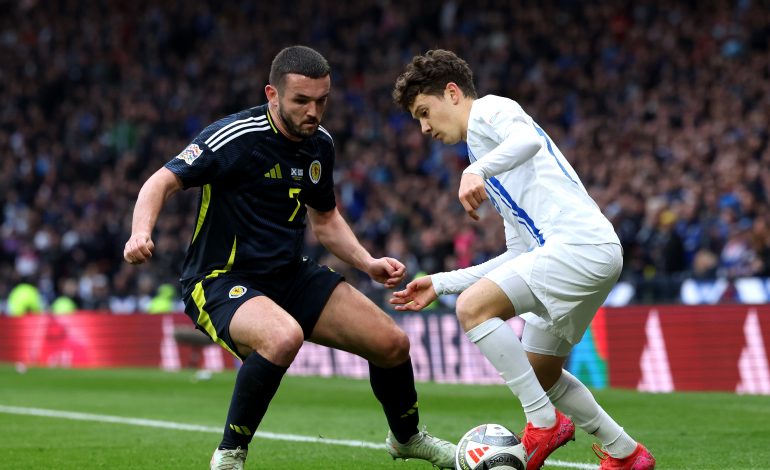 Zwei Spieler stehen sich gegenüber bei einem Fußballspiel. Der linke Spieler trägt ein schwarzes Trikot und spielt für Schottland. Rechts steht ein griechischer Nationalspieler in einem weißen Trikot. Der Griechische Spieler heißt Karetsas und versucht in diesem Moment an seinen Gegenspieler mit einem Dribbling vorbei zu kommen.