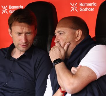 Simon Rolfes (Geschäftsführer Sport) und Fernando Carro (Vorsitzender der Geschäftsführung [CEO]) sitzen beide nebeneinander auf der Trainerbank in der BayArena. Fernando Carro hällt dabei seine linke Hand vor seinen Mund. Simon Rolfes hat seinen Knopf leicht nach unten gesenkt um näher an Carro zu sein. Beide haben einen ernsthaften Gesichtsausdruck.