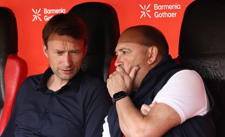 Simon Rolfes (Geschäftsführer Sport) und Fernando Carro (Vorsitzender der Geschäftsführung [CEO]) sitzen beide nebeneinander auf der Trainerbank in der BayArena. Fernando Carro hällt dabei seine linke Hand vor seinen Mund. Simon Rolfes hat seinen Knopf leicht nach unten gesenkt um näher an Carro zu sein. Beide haben einen ernsthaften Gesichtsausdruck.